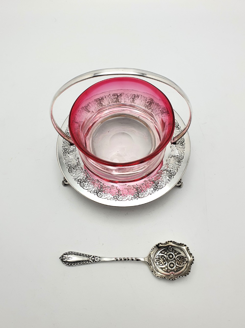 An Edwardian cranberry glass dish resting inside an engraved silver-plated stand with a swing handle and spoon. View V