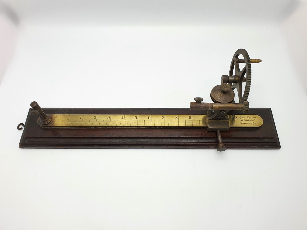 An antique brass thread tester mounted on a dark mahogany base with an engraved ruler scale and geared mechanism.