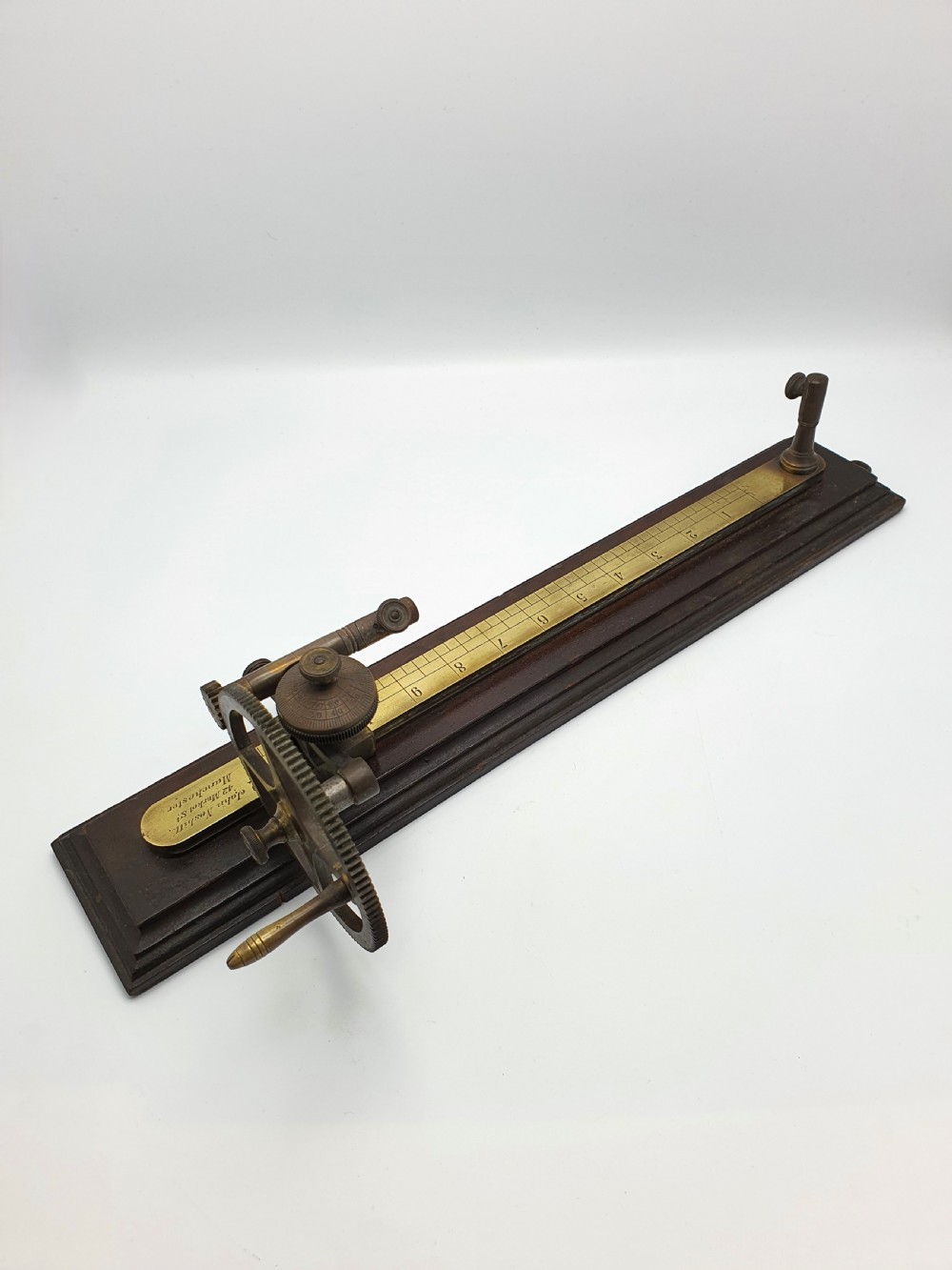 An antique brass thread tester mounted on a dark mahogany base with an engraved ruler scale and geared mechanism. View IV