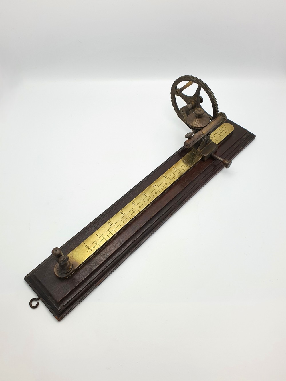 An antique brass thread tester mounted on a dark mahogany base with an engraved ruler scale and geared mechanism. View V