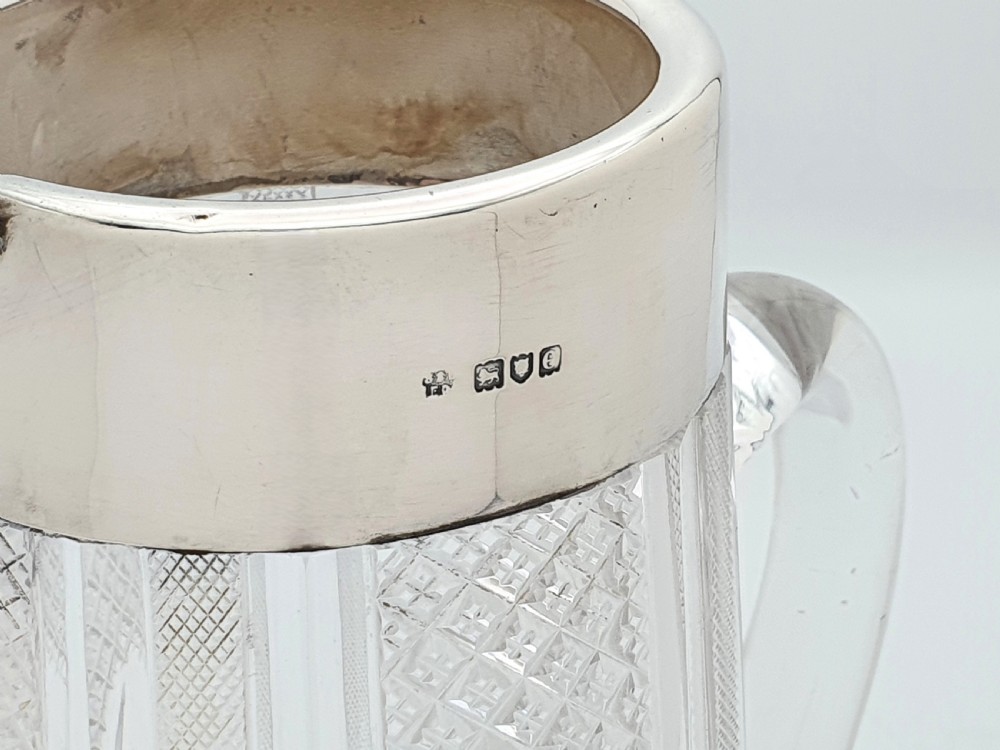 A late Victorian cut glass water jug featuring a tapered geometric cut body, an integrated glass handle, and a hallmarked sterling silver collar and spout. View VI