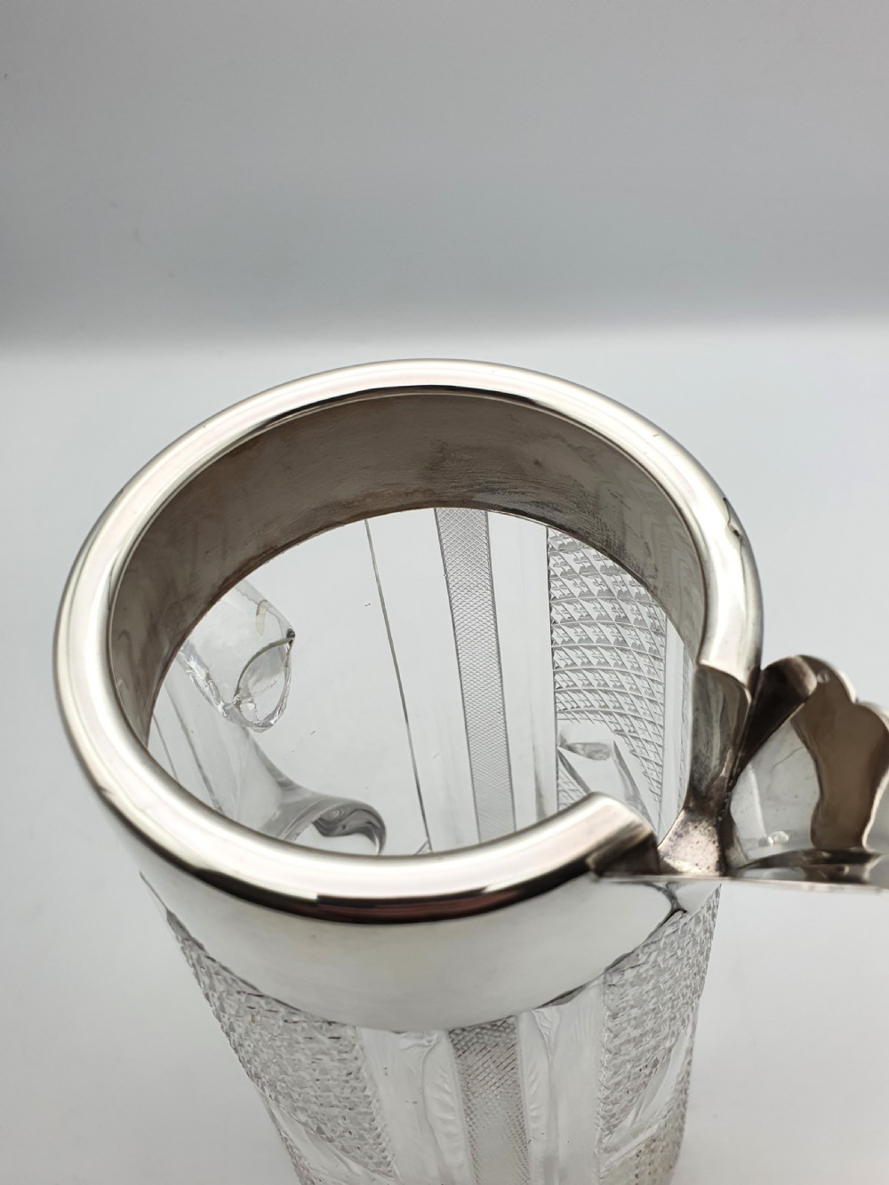 A late Victorian cut glass water jug featuring a tapered geometric cut body, an integrated glass handle, and a hallmarked sterling silver collar and spout. View IX
