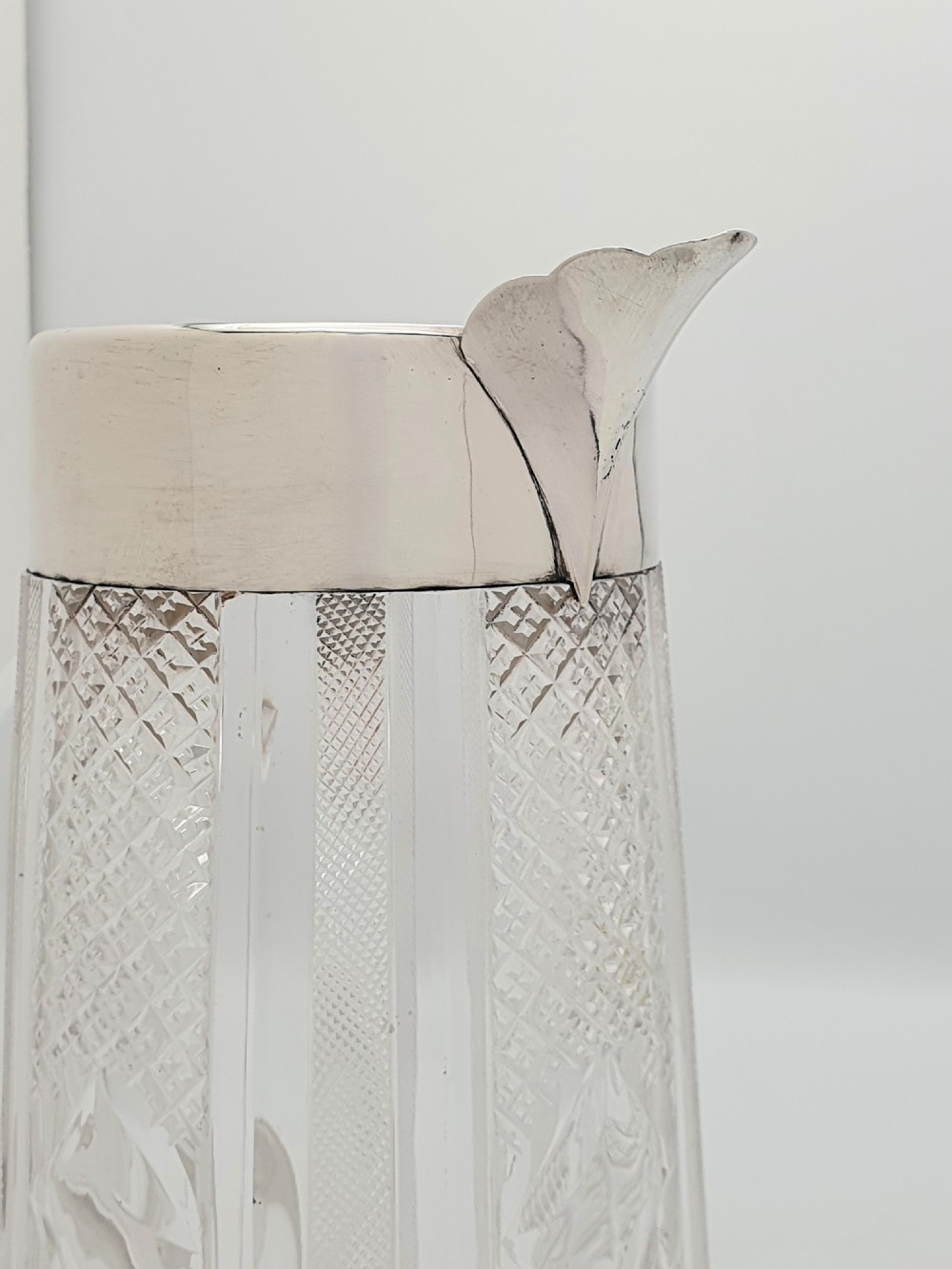 A late Victorian cut glass water jug featuring a tapered geometric cut body, an integrated glass handle, and a hallmarked sterling silver collar and spout. View X