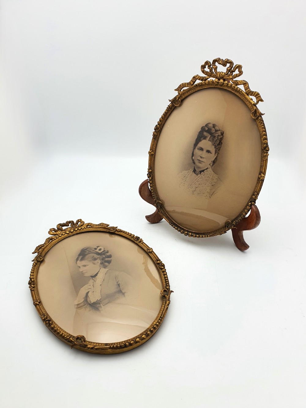 A pair of oval gilt brass photo frames featuring decorative ribbon bow crests and beaded borders containing sepia portrait photographs.