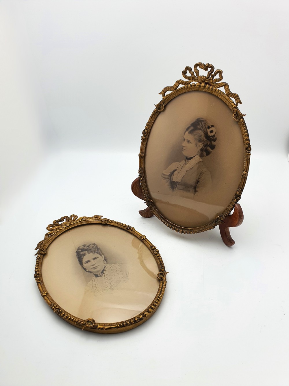 A pair of oval gilt brass photo frames featuring decorative ribbon bow crests and beaded borders containing sepia portrait photographs. View II