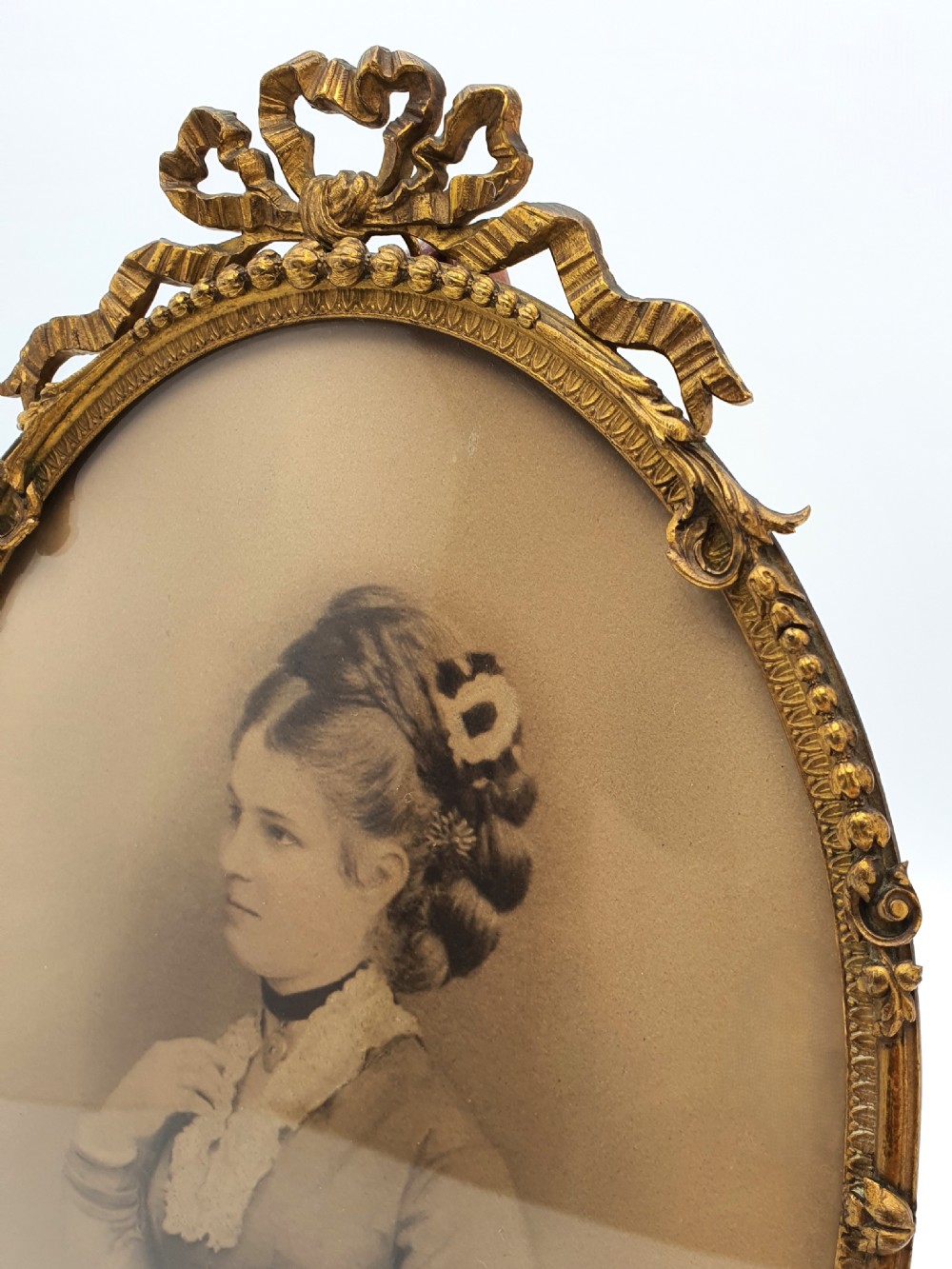 A pair of oval gilt brass photo frames featuring decorative ribbon bow crests and beaded borders containing sepia portrait photographs. View V