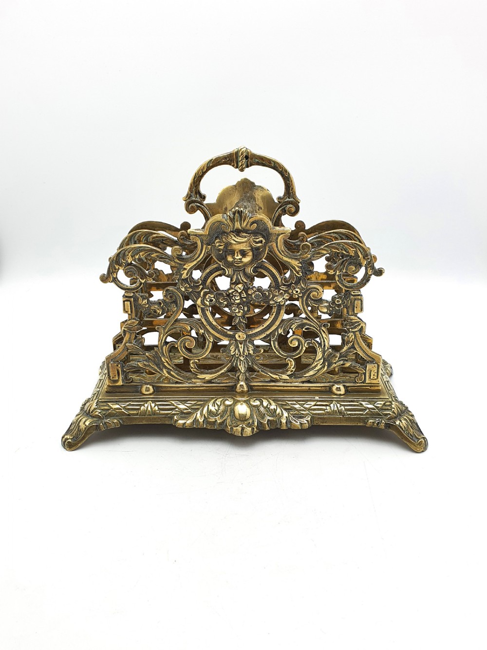 An ornate cast brass letter rack featuring openwork scrollwork, a central cherub face motif, a top handle, and a decorated rectangular base.