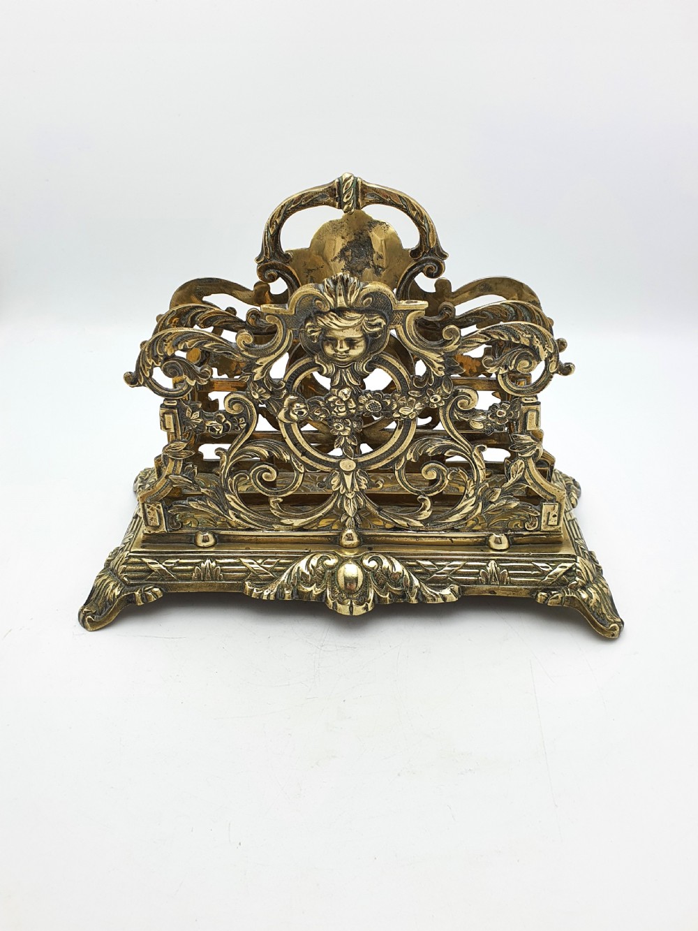An ornate cast brass letter rack featuring openwork scrollwork, a central cherub face motif, a top handle, and a decorated rectangular base. View III