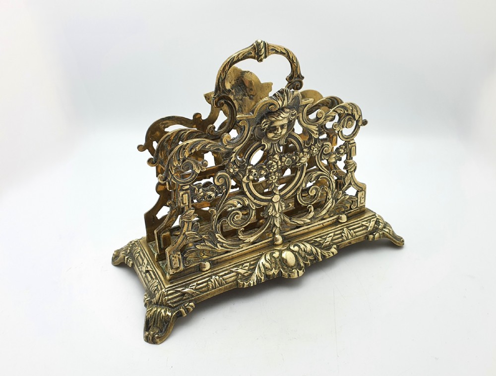 An ornate cast brass letter rack featuring openwork scrollwork, a central cherub face motif, a top handle, and a decorated rectangular base. View V