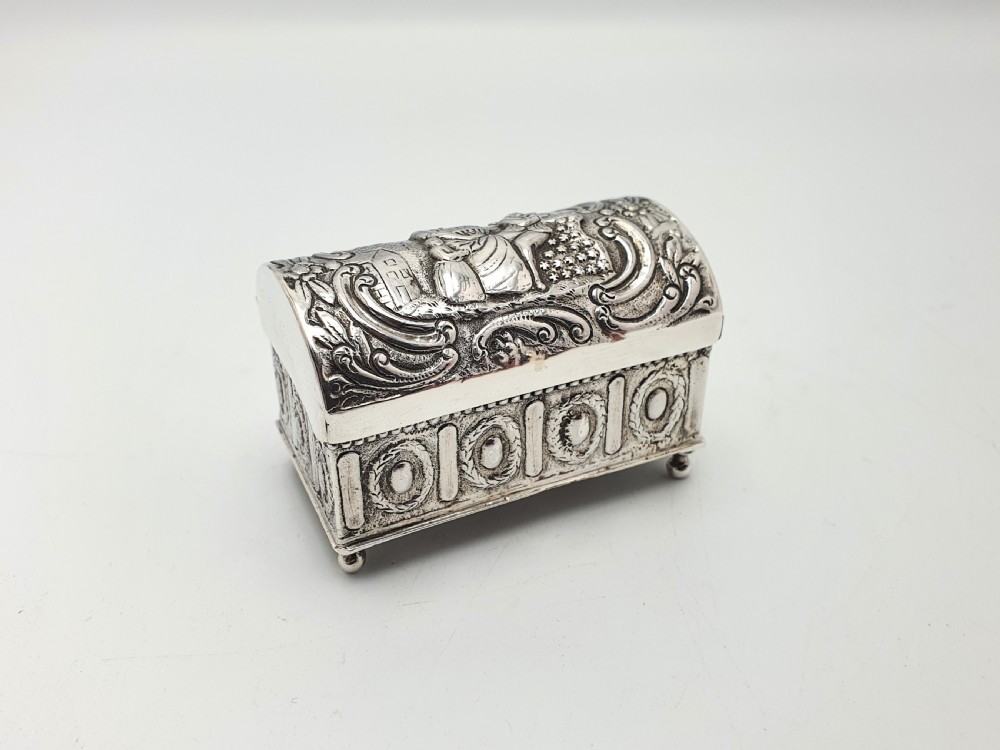 An embossed silver rectangular trinket box featuring a domed lid decorated with figures in relief and supported by four small ball feet.