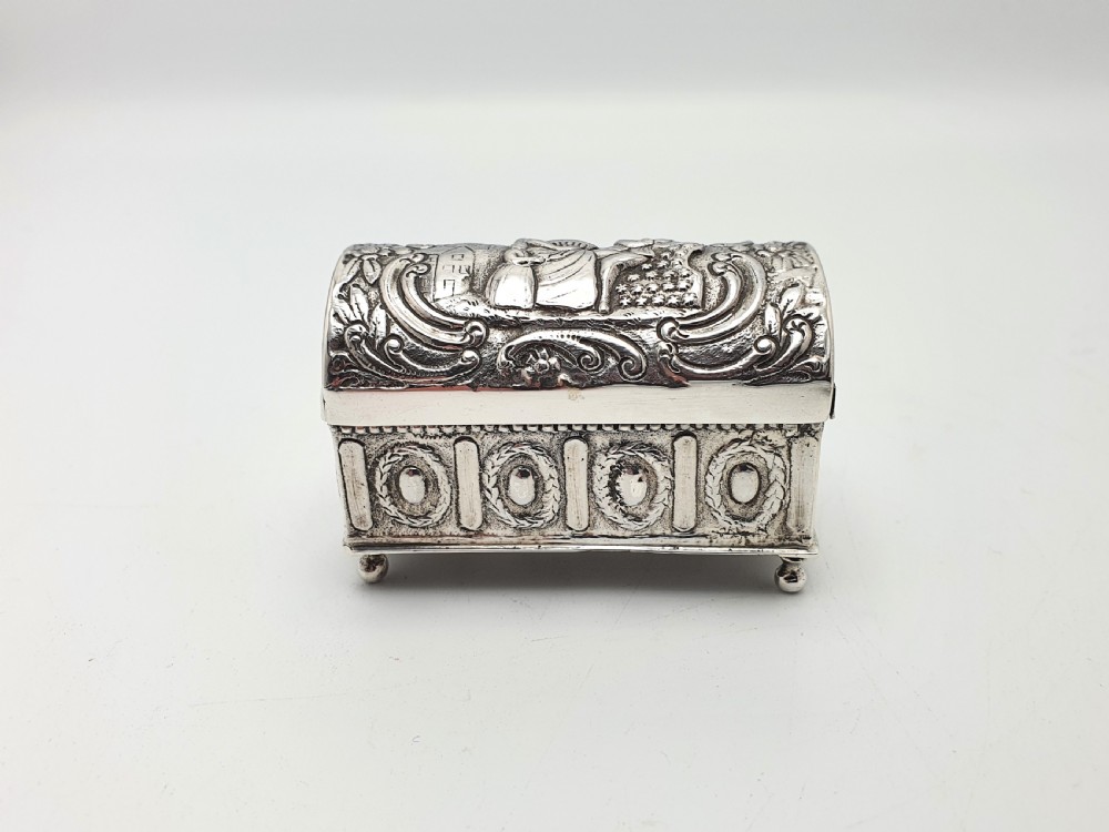An embossed silver rectangular trinket box featuring a domed lid decorated with figures in relief and supported by four small ball feet. View II