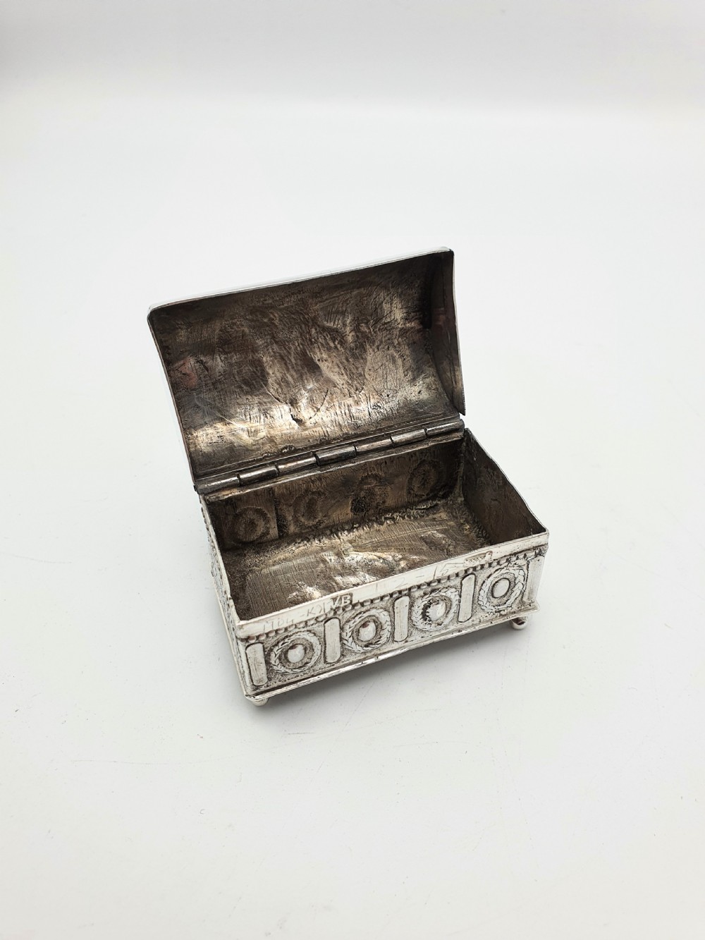 An embossed silver rectangular trinket box featuring a domed lid decorated with figures in relief and supported by four small ball feet. View III