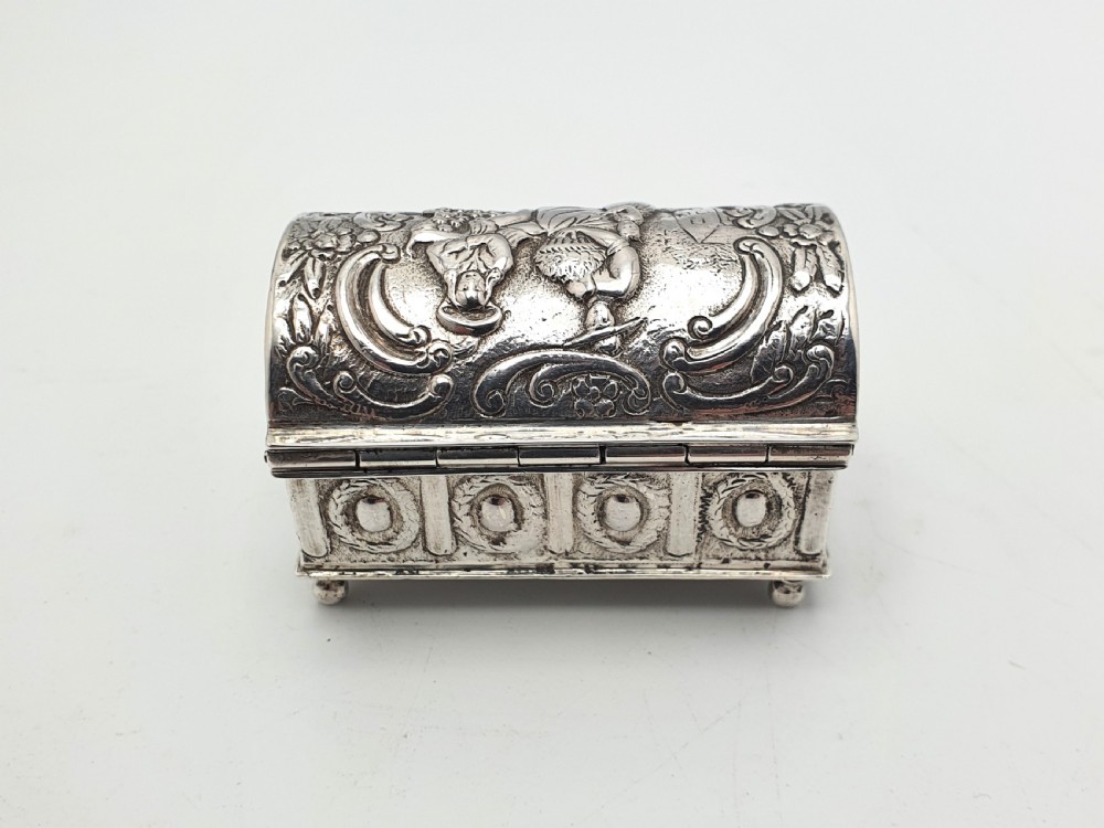 An embossed silver rectangular trinket box featuring a domed lid decorated with figures in relief and supported by four small ball feet. View VII