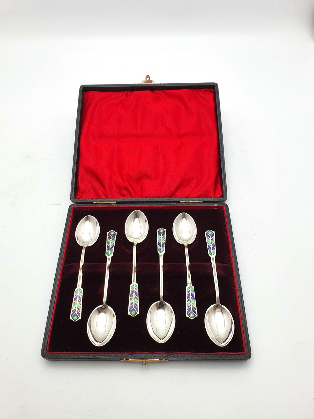 A set of six sterling silver teaspoons featuring green and blue geometric enamelled handles presented inside a fitted black velvet case with a red silk lid lining.