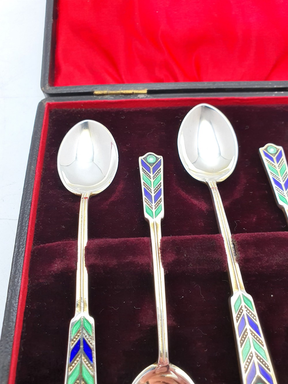 A set of six sterling silver teaspoons featuring green and blue geometric enamelled handles presented inside a fitted black velvet case with a red silk lid lining. View II