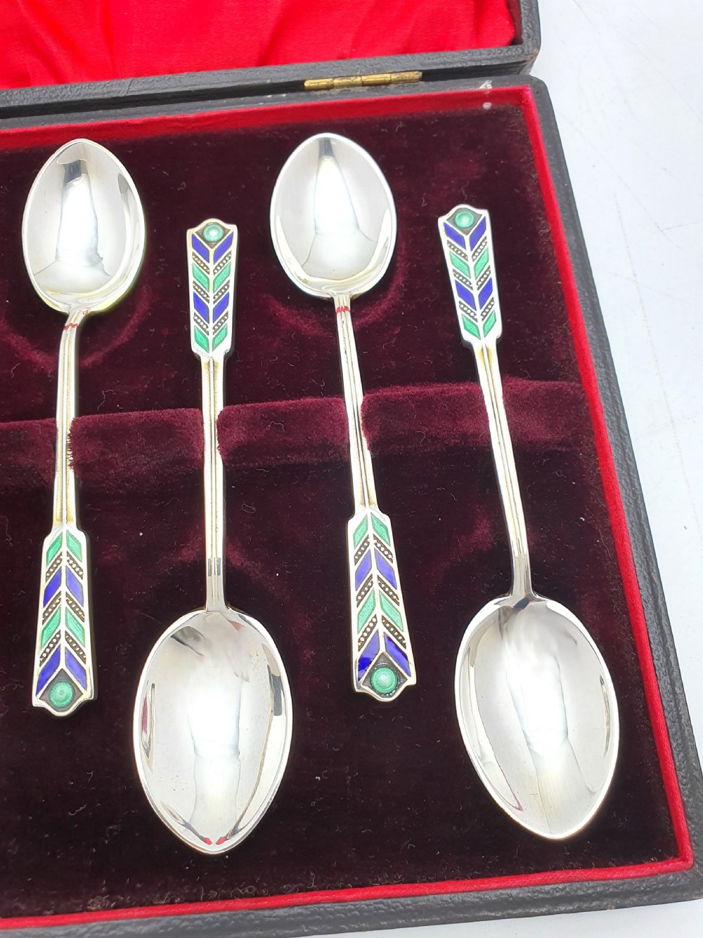 A set of six sterling silver teaspoons featuring green and blue geometric enamelled handles presented inside a fitted black velvet case with a red silk lid lining. View III