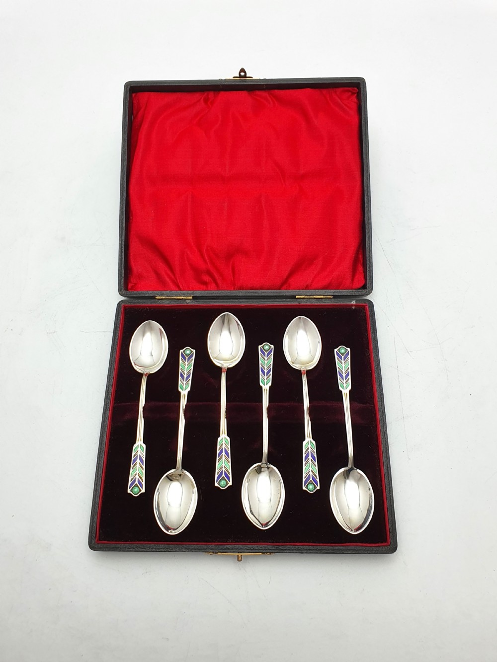 A set of six sterling silver teaspoons featuring green and blue geometric enamelled handles presented inside a fitted black velvet case with a red silk lid lining. View IV