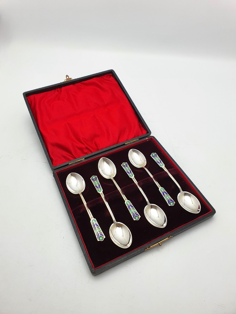 A set of six sterling silver teaspoons featuring green and blue geometric enamelled handles presented inside a fitted black velvet case with a red silk lid lining. View VII
