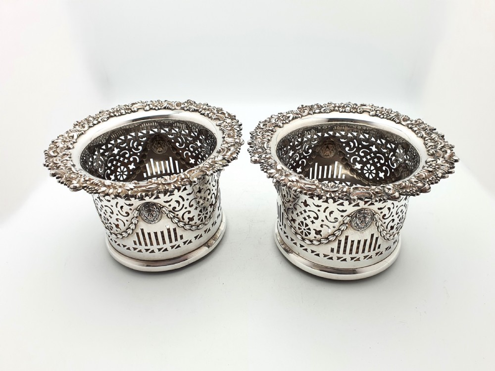 A pair of circular silver-plated wine coasters with pierced fretwork sides, garland swags, medallion motifs, and decorative rims.