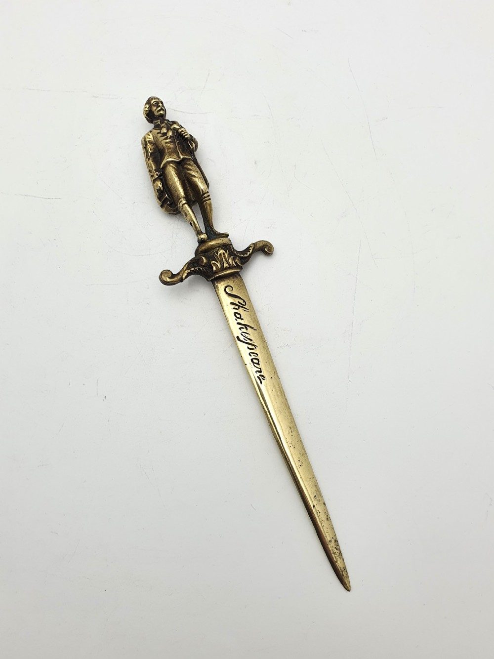 An antique brass letter opener shaped like a dagger featuring a sculpted figure of William Shakespeare on the handle and his name engraved on the blade.