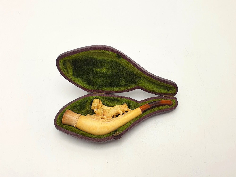 An antique carved meerschaum cheroot holder featuring detailed hunting dogs resting inside a green velvet-lined fitted case.
