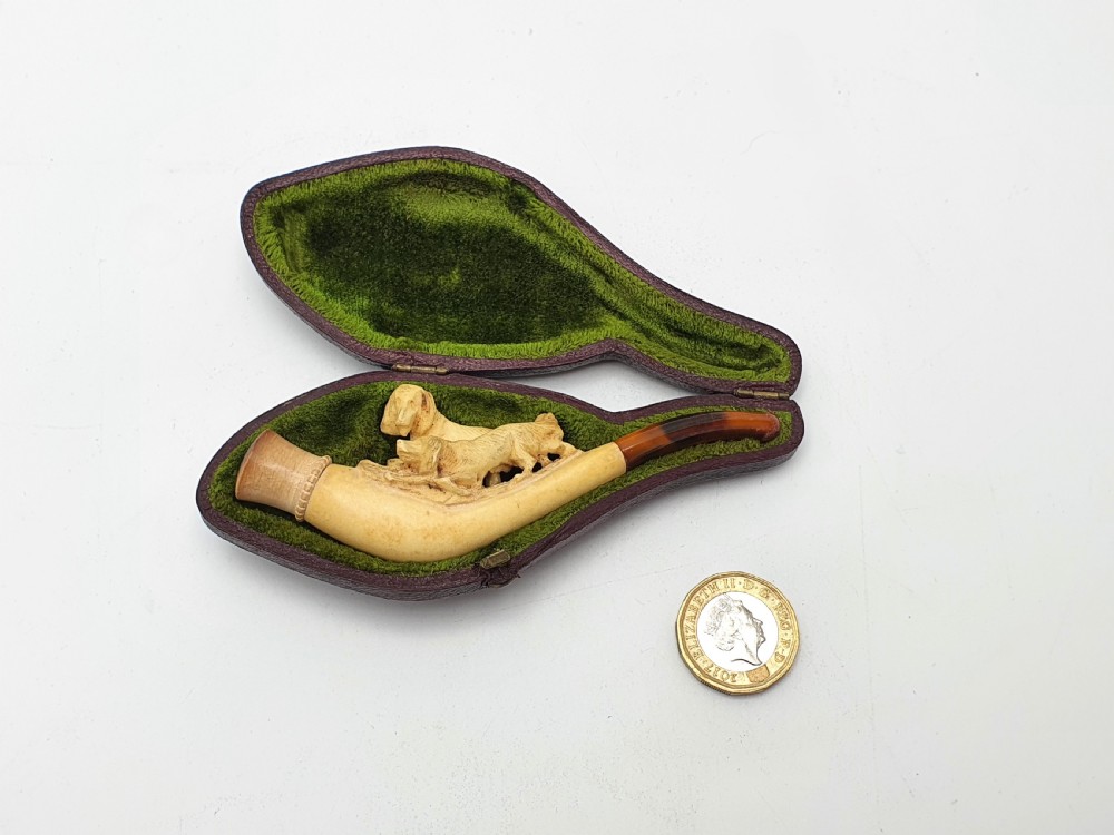 An antique carved meerschaum cheroot holder featuring detailed hunting dogs resting inside a green velvet-lined fitted case. View V