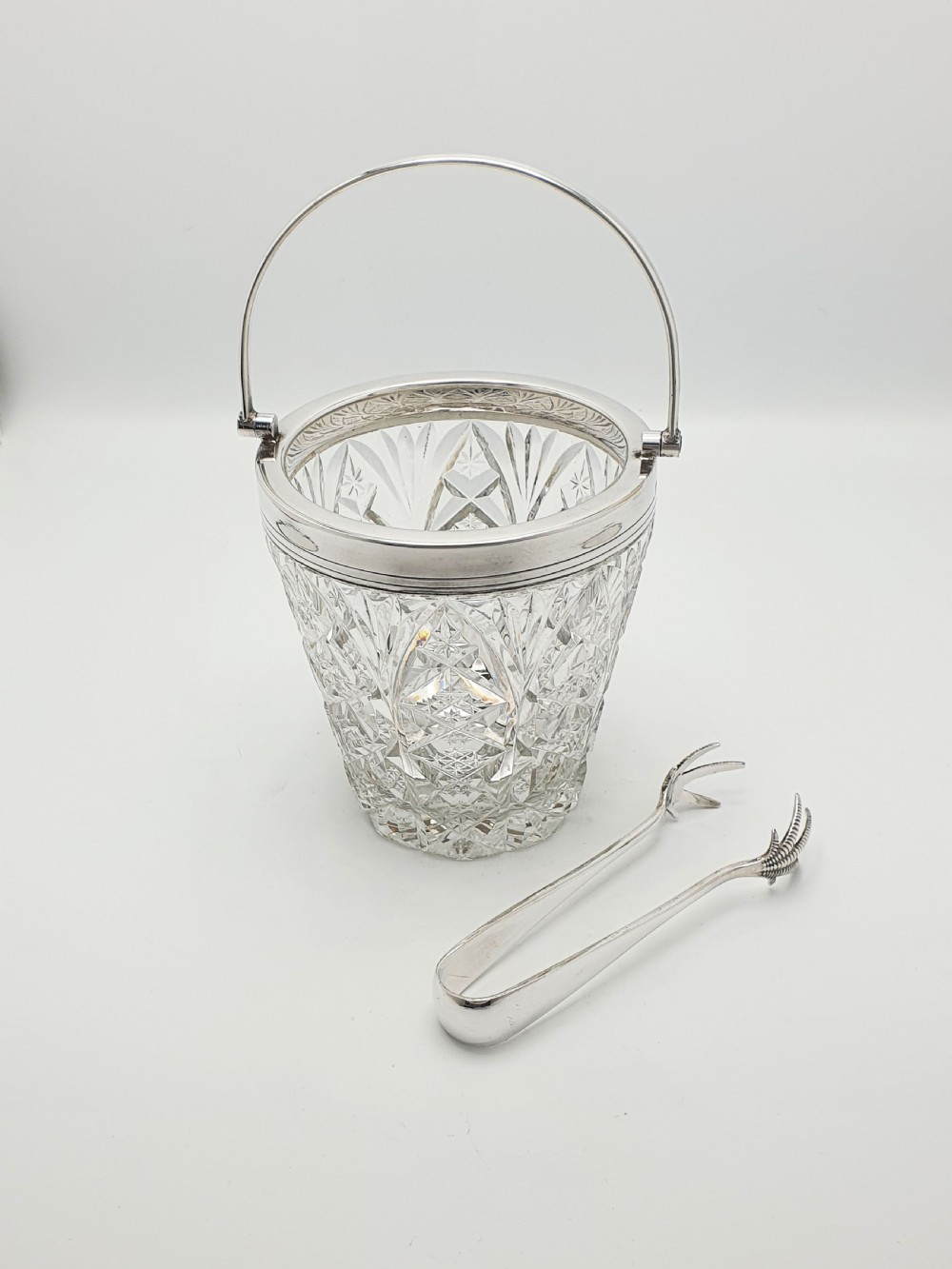 A cut glass ice bucket with a silver-plated rim and carrying handle, resting beside a pair of silver-plated ice tongs with claw tips.