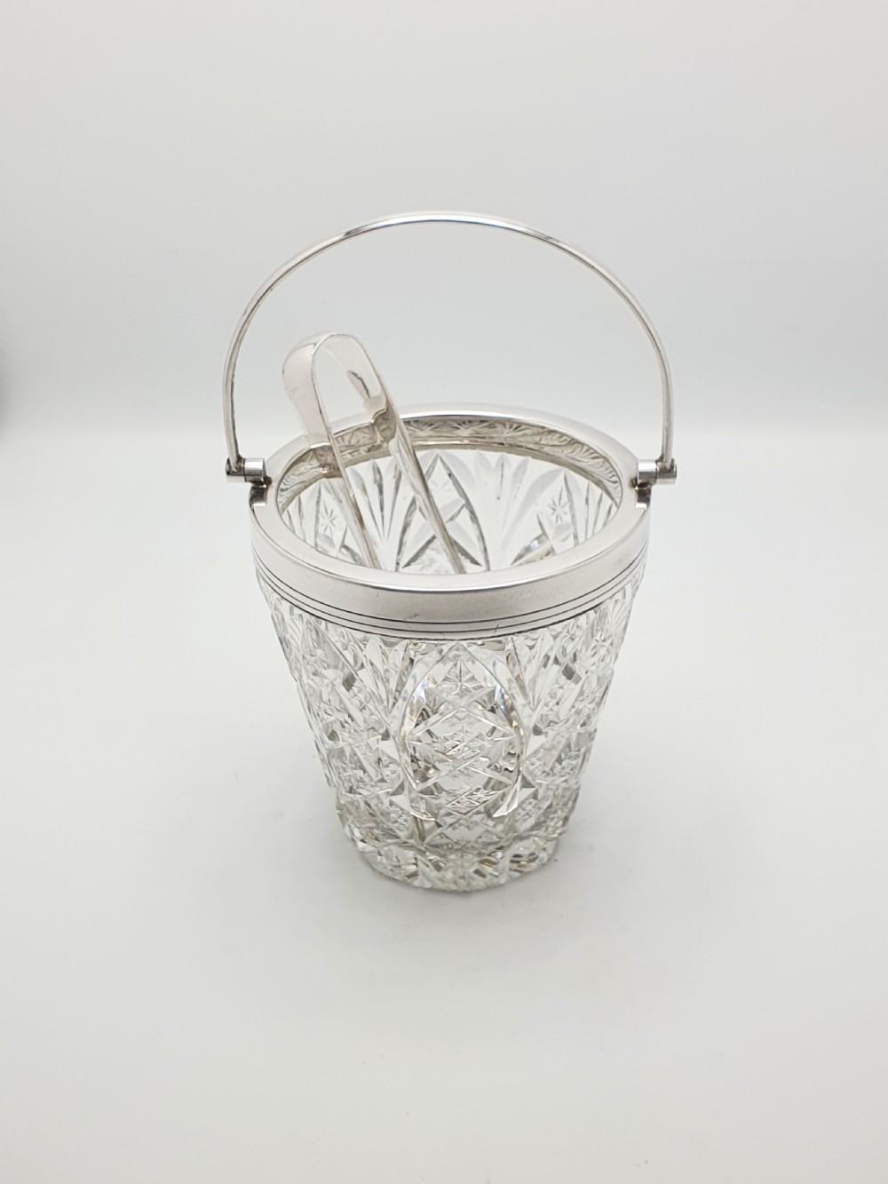 A cut glass ice bucket with a silver-plated rim and carrying handle, resting beside a pair of silver-plated ice tongs with claw tips. View II