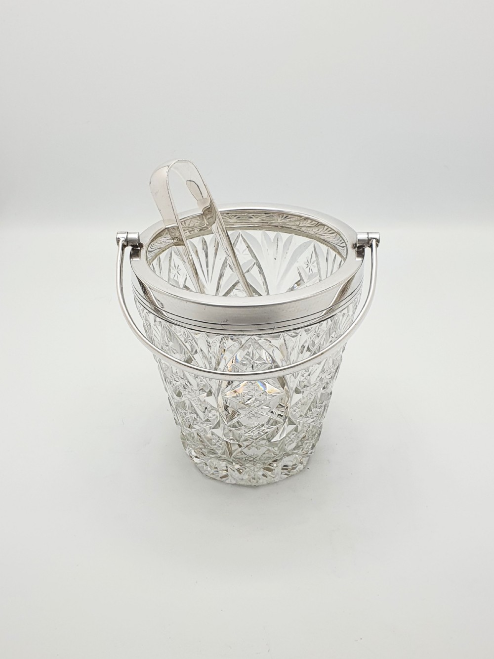 A cut glass ice bucket with a silver-plated rim and carrying handle, resting beside a pair of silver-plated ice tongs with claw tips. View III