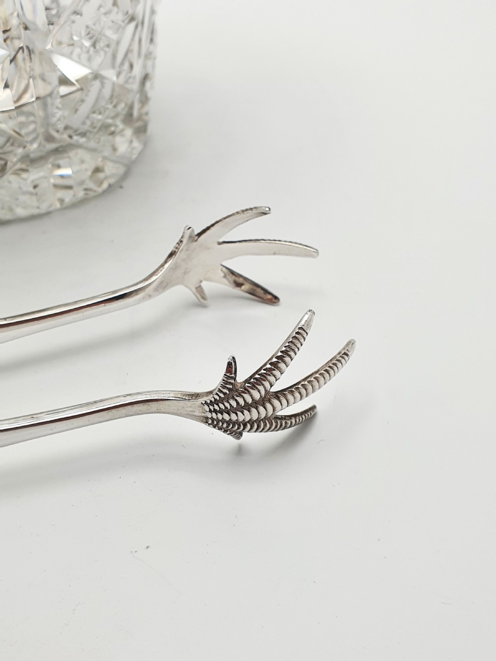 A cut glass ice bucket with a silver-plated rim and carrying handle, resting beside a pair of silver-plated ice tongs with claw tips. View V