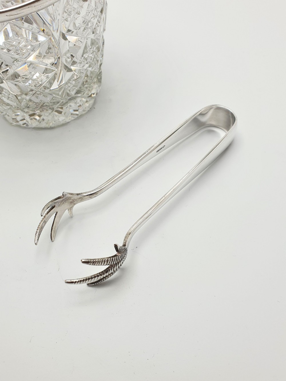 A cut glass ice bucket with a silver-plated rim and carrying handle, resting beside a pair of silver-plated ice tongs with claw tips. View VI