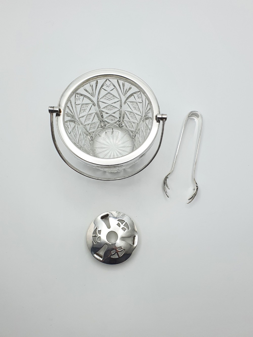 A cut glass ice bucket with a silver-plated rim and carrying handle, resting beside a pair of silver-plated ice tongs with claw tips. View VII