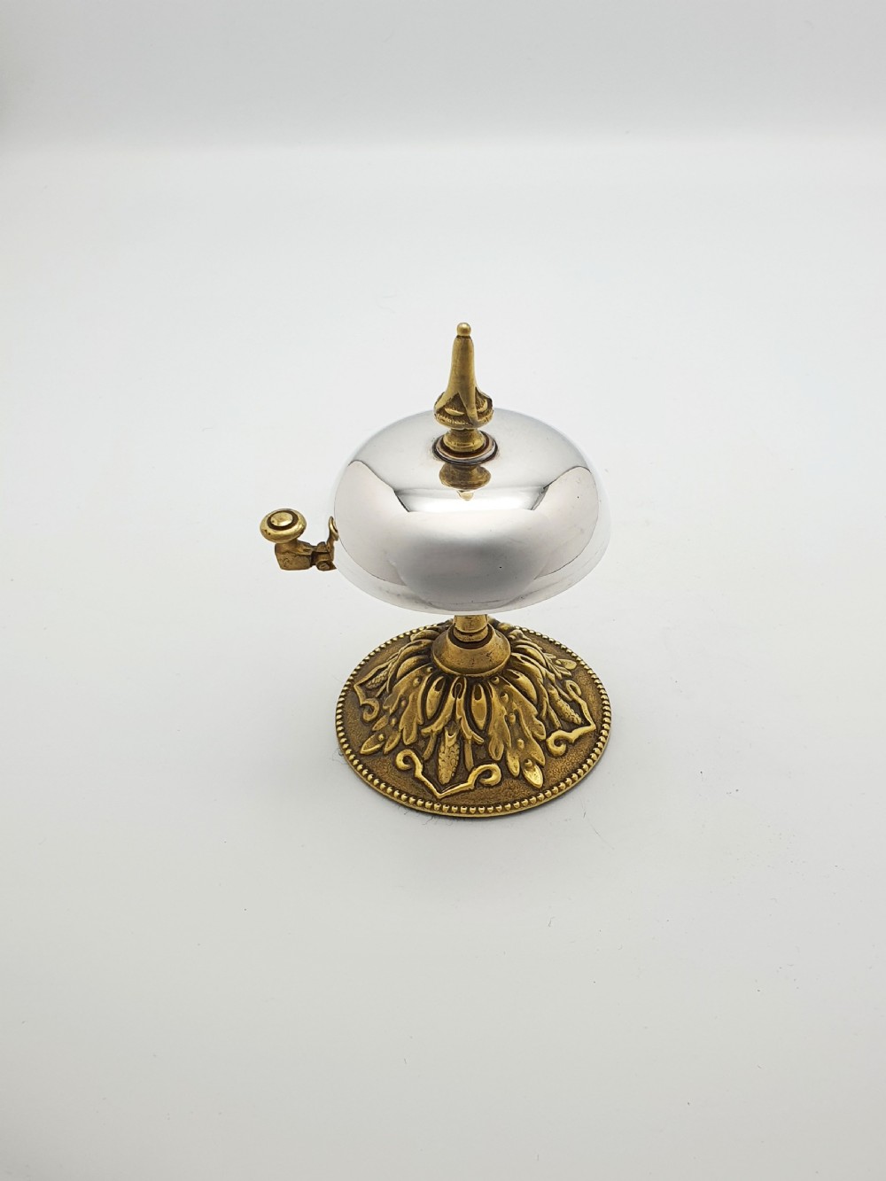 A silver-plated dome desk bell with a brass plunger mechanism mounted on an ornate circular brass base decorated with acanthus leaf motifs.