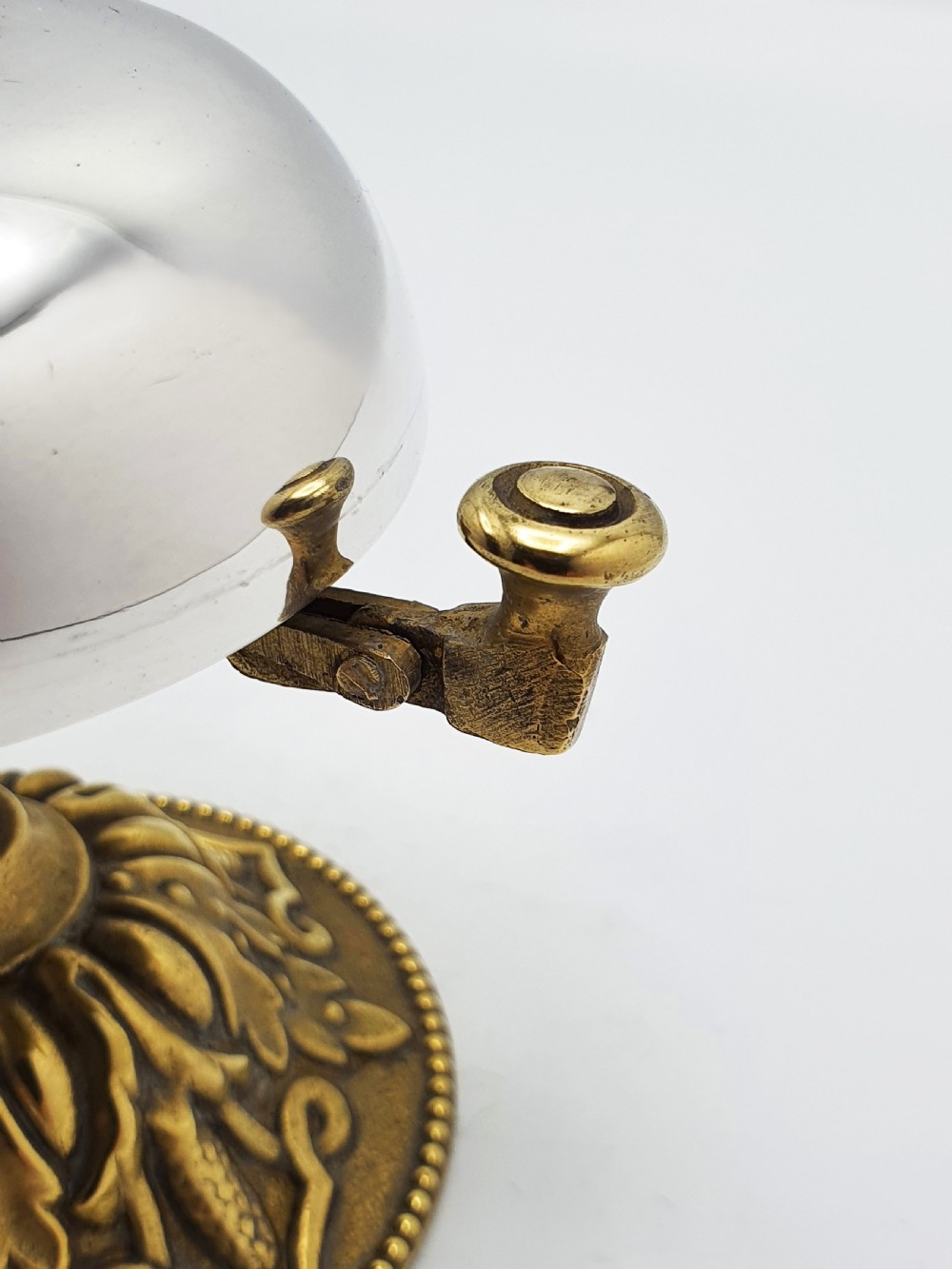 A silver-plated dome desk bell with a brass plunger mechanism mounted on an ornate circular brass base decorated with acanthus leaf motifs. View V