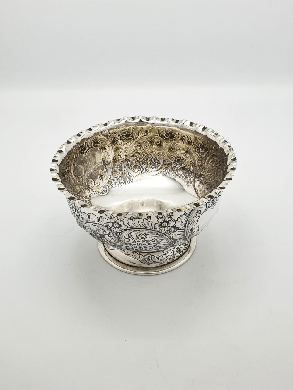 A circular Victorian sterling silver bowl featuring ornate repousse floral chasing, a crimped fluted rim, and a raised pedestal foot.