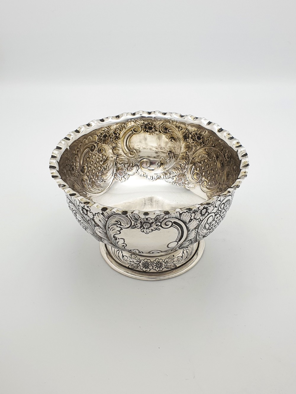 A circular Victorian sterling silver bowl featuring ornate repousse floral chasing, a crimped fluted rim, and a raised pedestal foot. View II