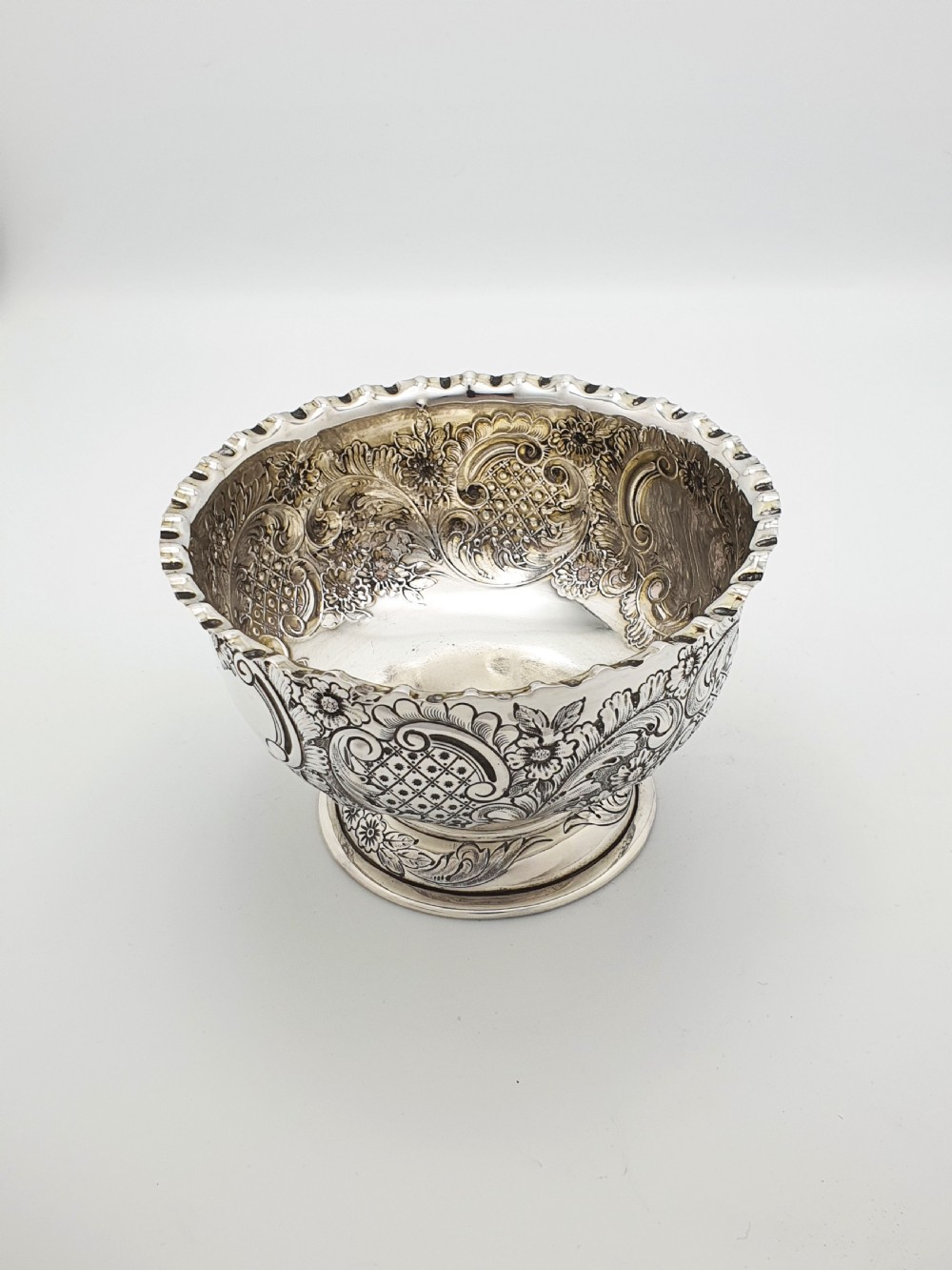A circular Victorian sterling silver bowl featuring ornate repousse floral chasing, a crimped fluted rim, and a raised pedestal foot. View III