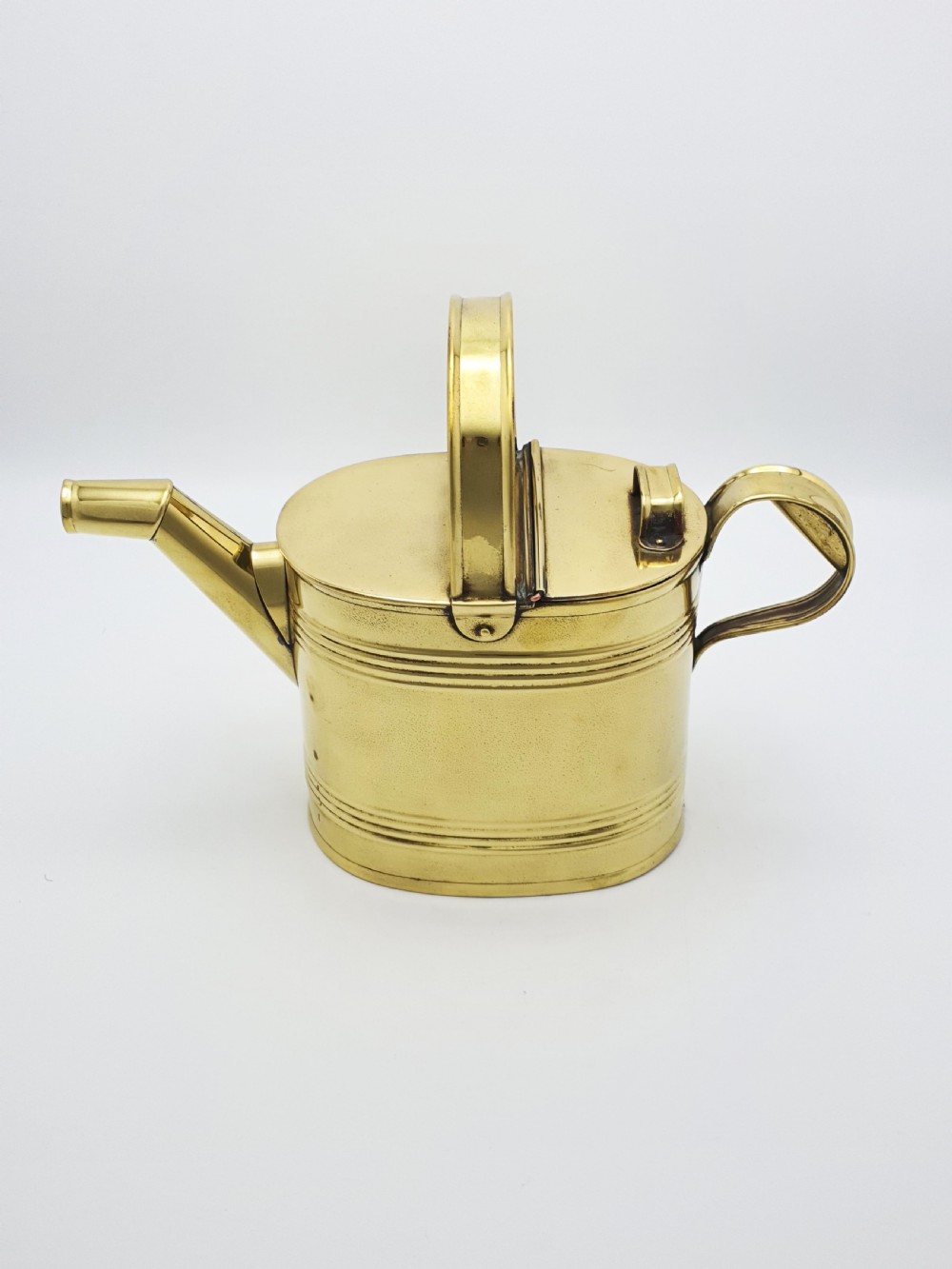 A polished late Victorian brass watering can with an oval body, a curved upper bridge handle, a rear loop handle, and a straight tapered spout. View II
