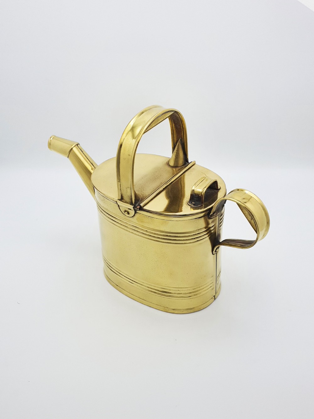 A polished late Victorian brass watering can with an oval body, a curved upper bridge handle, a rear loop handle, and a straight tapered spout. View VI