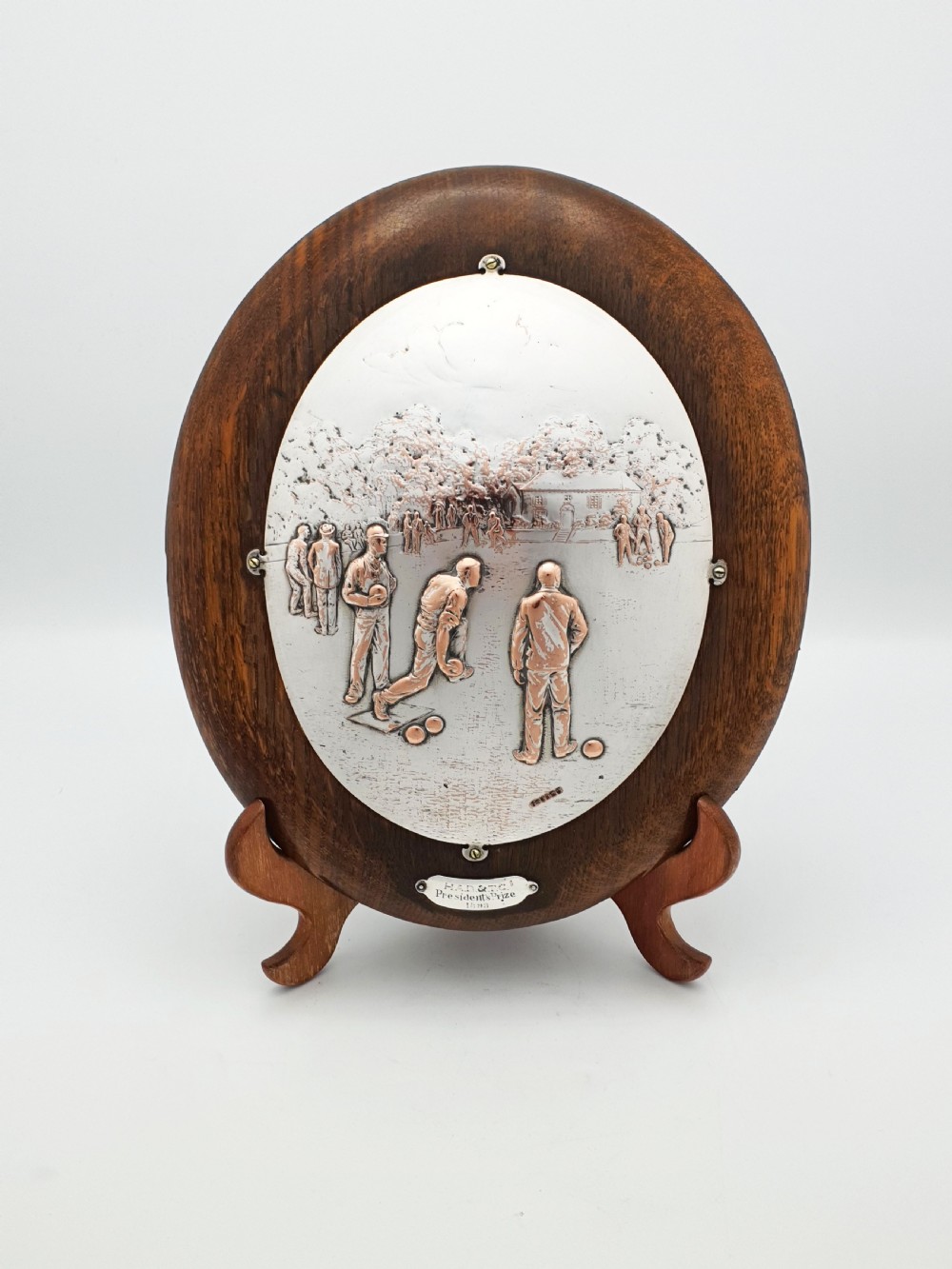 An oval timber plaque displaying an embossed silver-plated metal relief depiction of late Victorian lawn bowling players dated 1899.