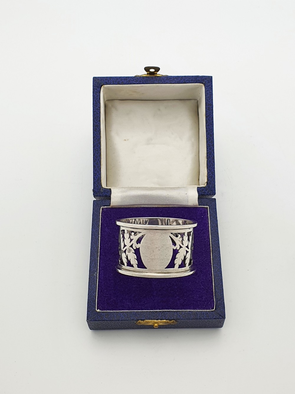An Edwardian sterling silver napkin ring featuring intricate pierced leaf motifs and an unengraved central oval cartouche, presented in a velvet-lined blue box.