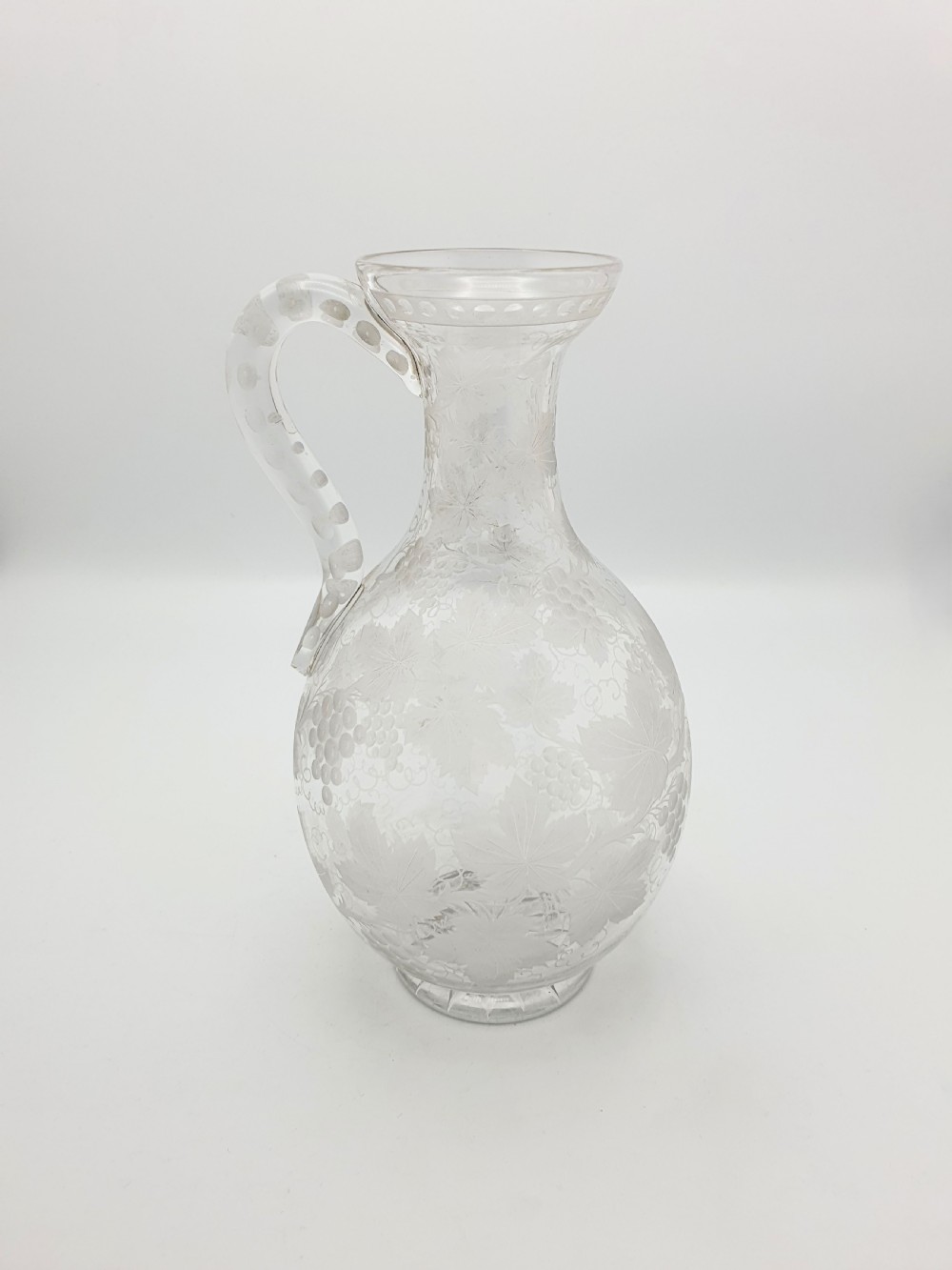 A late 19th-century clear glass wine jug decorated with etched grapevine motifs, a flared rim, and a curved applied handle.