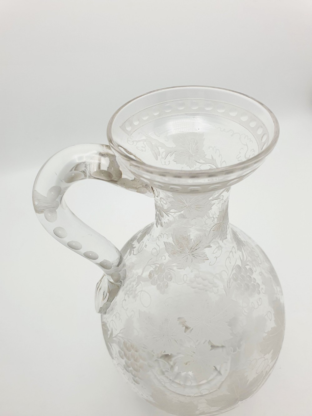 A late 19th-century clear glass wine jug decorated with etched grapevine motifs, a flared rim, and a curved applied handle. View VI
