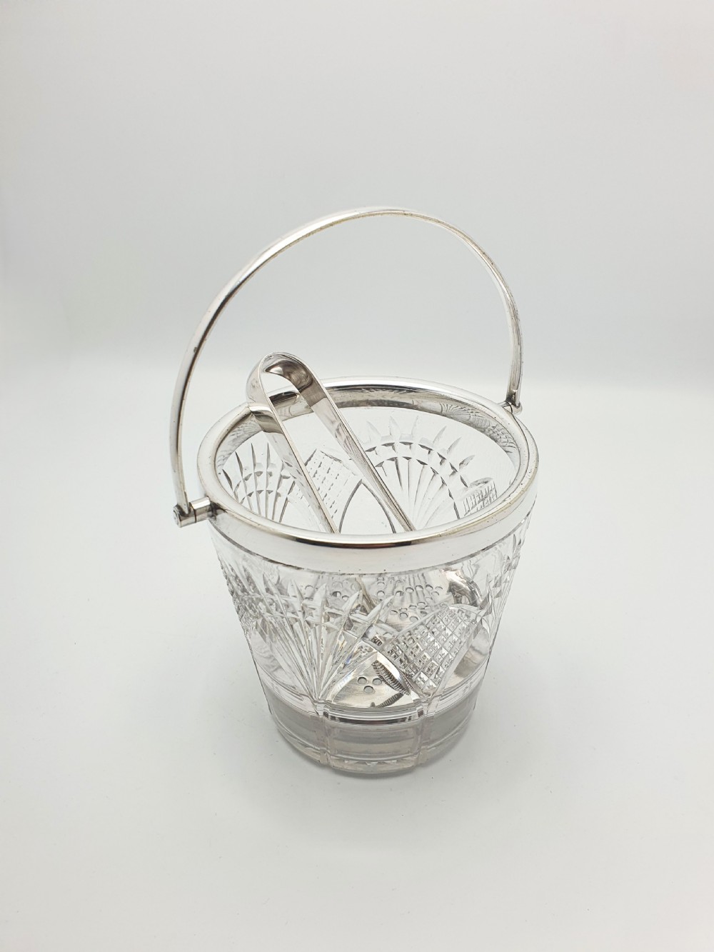 A clear cut-glass ice bucket featuring a silver-plated rim, swing handle, internal strainer plate, and matching metal ice tongs.