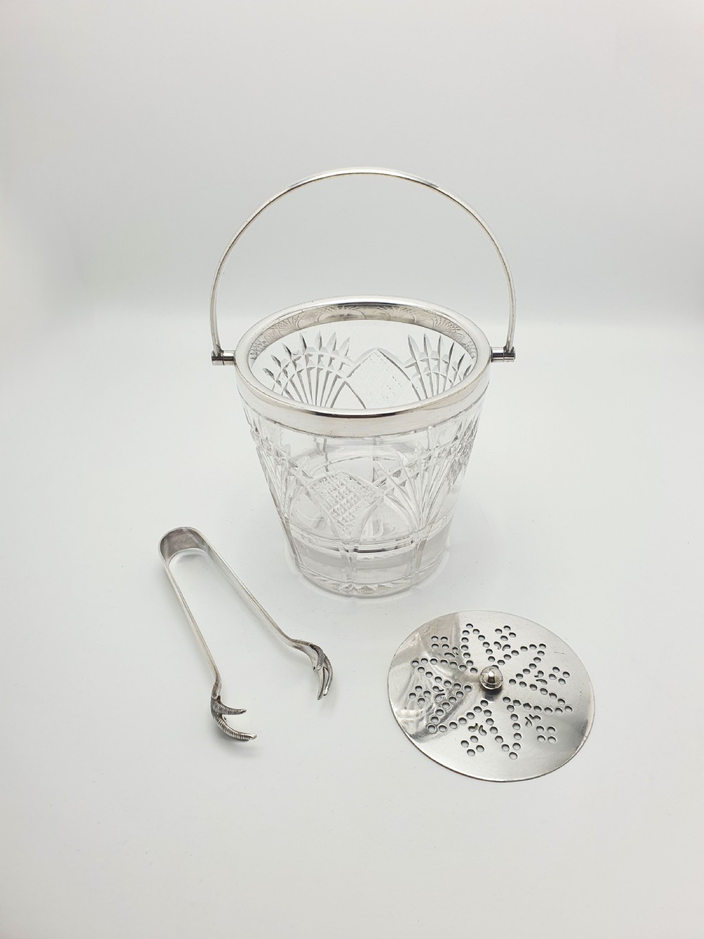 A clear cut-glass ice bucket featuring a silver-plated rim, swing handle, internal strainer plate, and matching metal ice tongs. View II