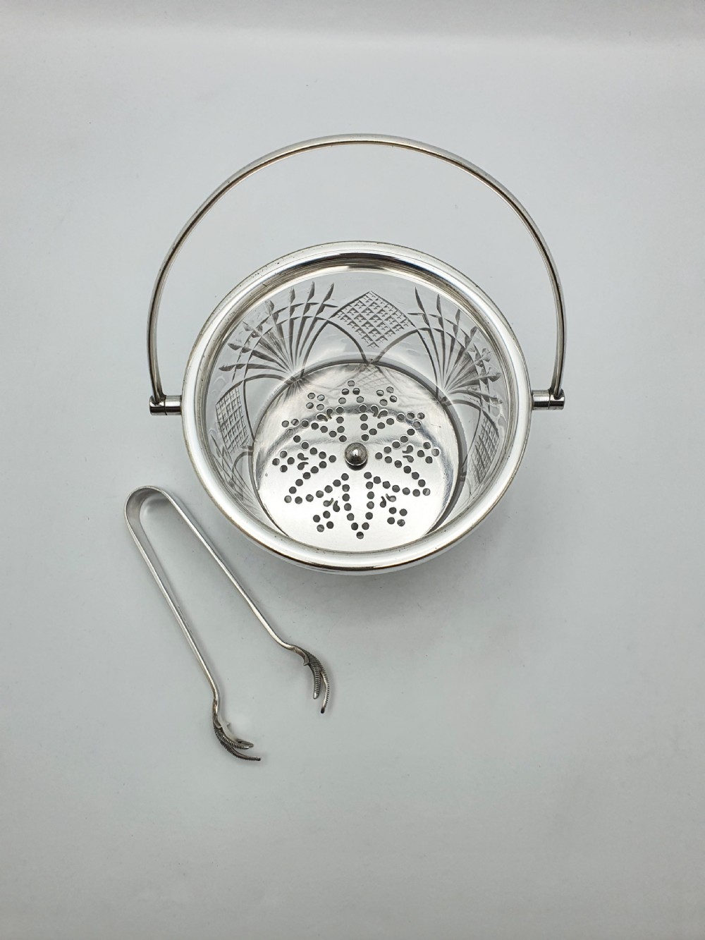A clear cut-glass ice bucket featuring a silver-plated rim, swing handle, internal strainer plate, and matching metal ice tongs. View III