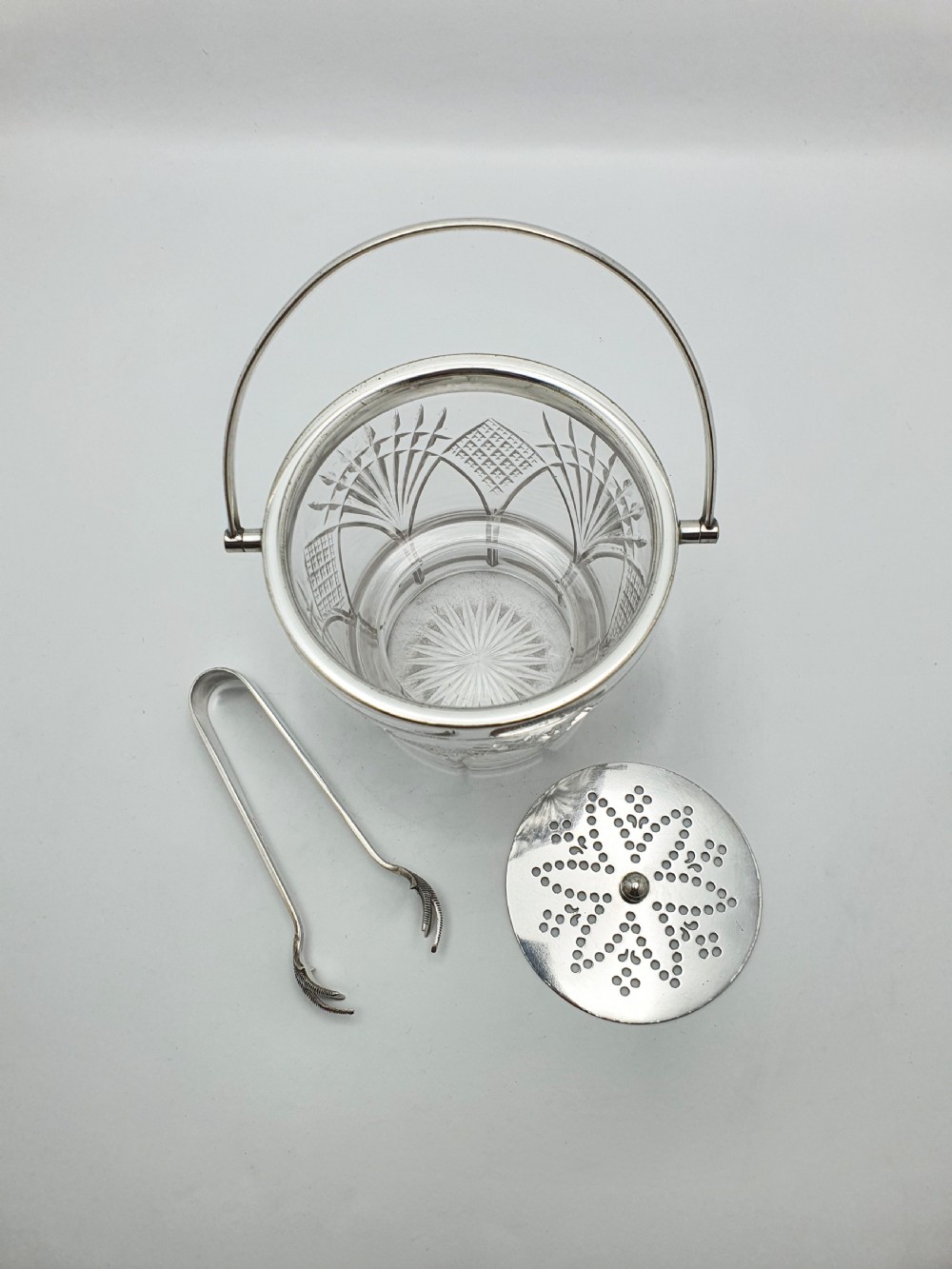 A clear cut-glass ice bucket featuring a silver-plated rim, swing handle, internal strainer plate, and matching metal ice tongs. View V