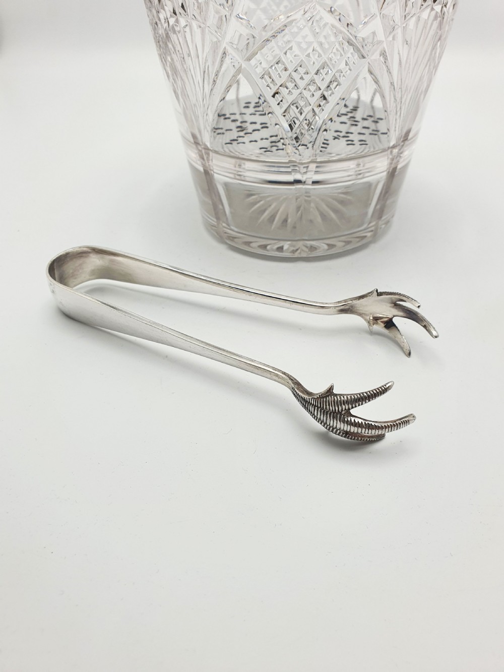 A clear cut-glass ice bucket featuring a silver-plated rim, swing handle, internal strainer plate, and matching metal ice tongs. View VI
