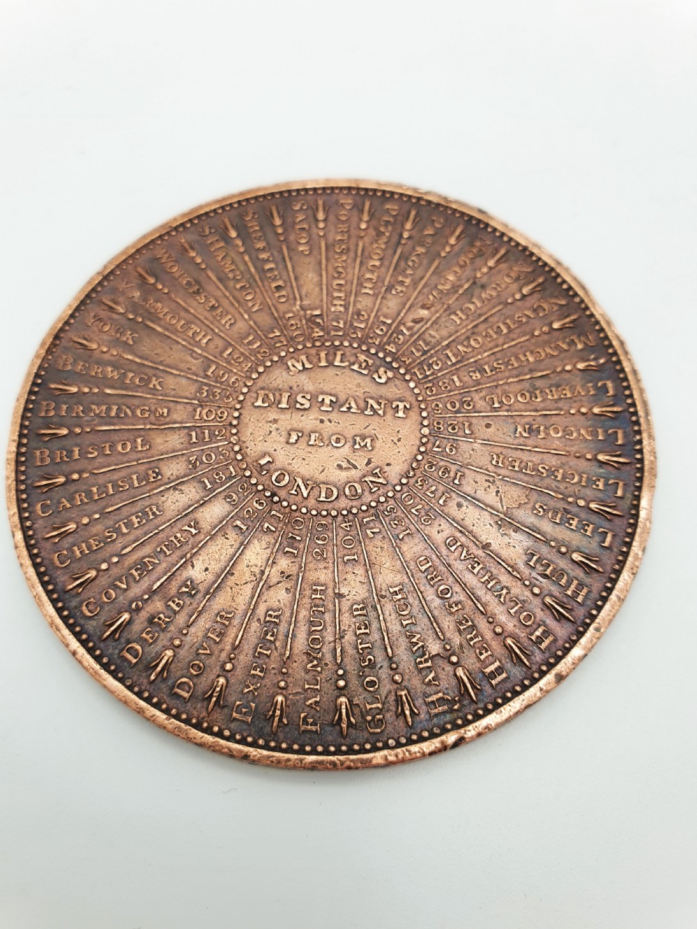 A circular patinated bronze medallion featuring radiating text detailing distances in miles from London to various British towns and cities.