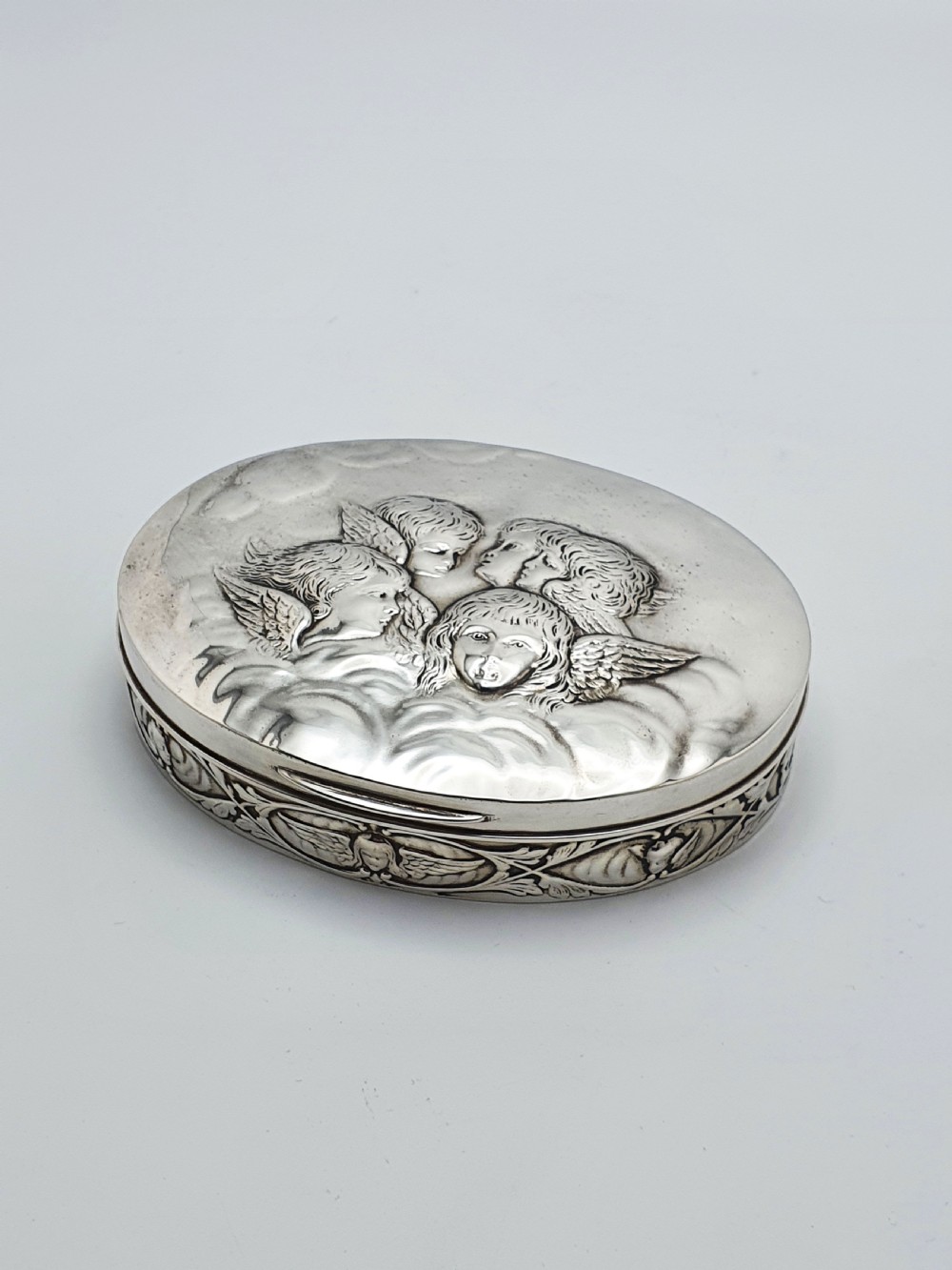 An oval sterling silver trinket box featuring an embossed lid with four cherub heads and a foliate border along the rim.